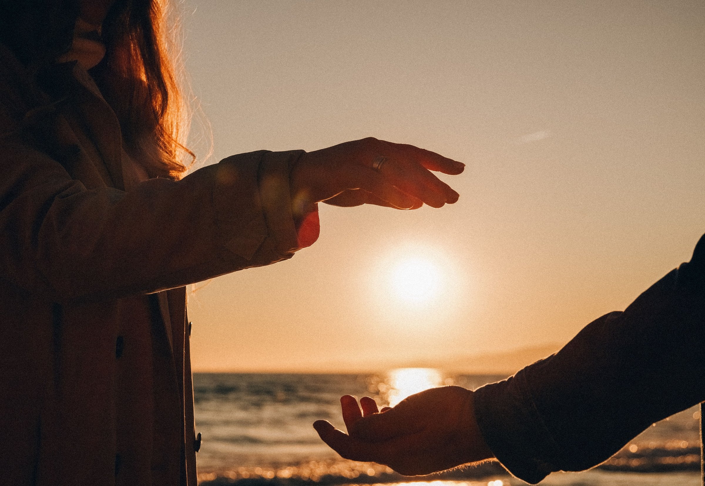 Deux mains qui se tendent l’une vers l’autre devant un coucher de soleil sur la mer.